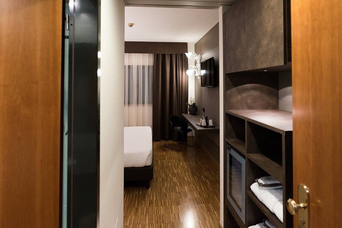 A hotel room with a bed, a desk, a chair, a TV, and a shelf with towels.