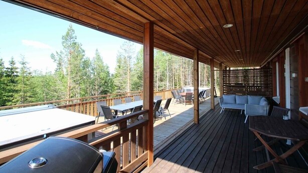 Eine überdachte Terrasse im Freien mit Holzboden, Tischen, Stühlen und einem Grill.
