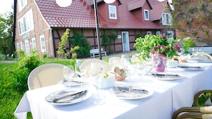 An outdoor dining setup with a table set for a meal, surrounded by greenery and a brick building in the background. - Landhaus Elbeflair Apartment GmbH (Lenzerwische)