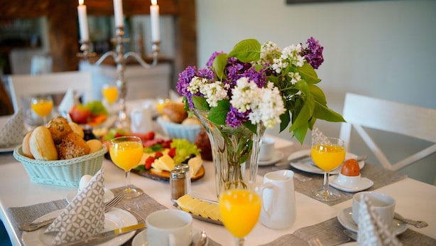 A breakfast setting with a table set for two, including plates, cutlery, glasses of orange juice, a vase with flowers, and a basket of bread. - Landhaus Elbeflair Apartment GmbH (Lenzerwische)