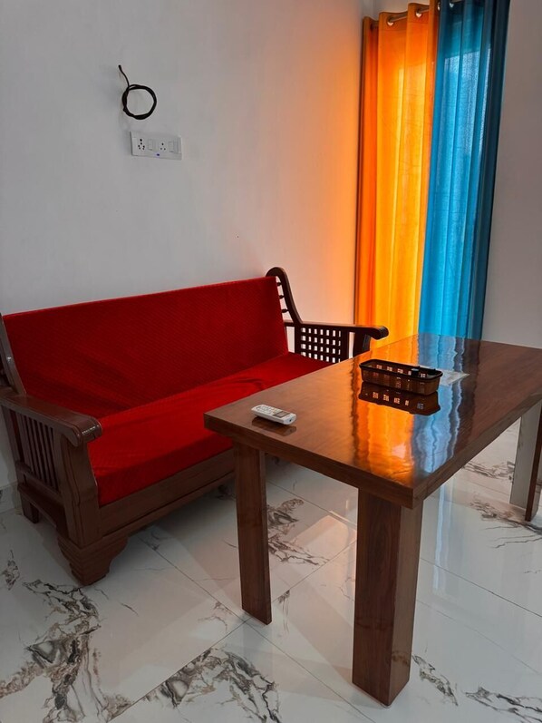 A room with a red sofa, a wooden table, a remote control, and a power outlet on the wall. - Ramalayam (Faizabad)