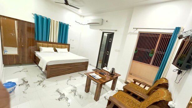 A hotel room with a bed, a wooden table, a sofa, a wardrobe, a ceiling fan, and a window with curtains. - Ramalayam (Faizabad)