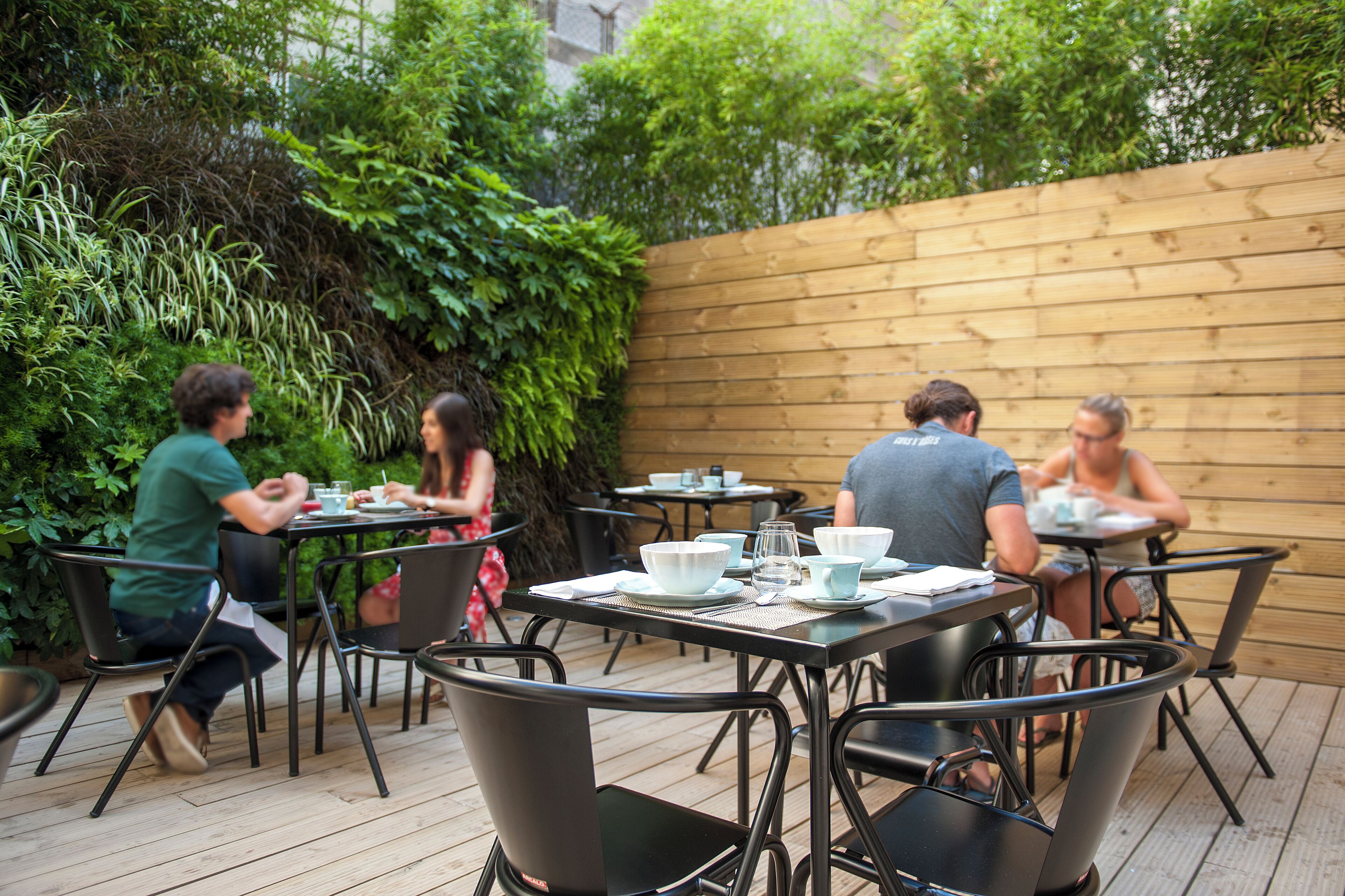 An outdoor patio with tables and chairs, surrounded by greenery and a wooden fence.