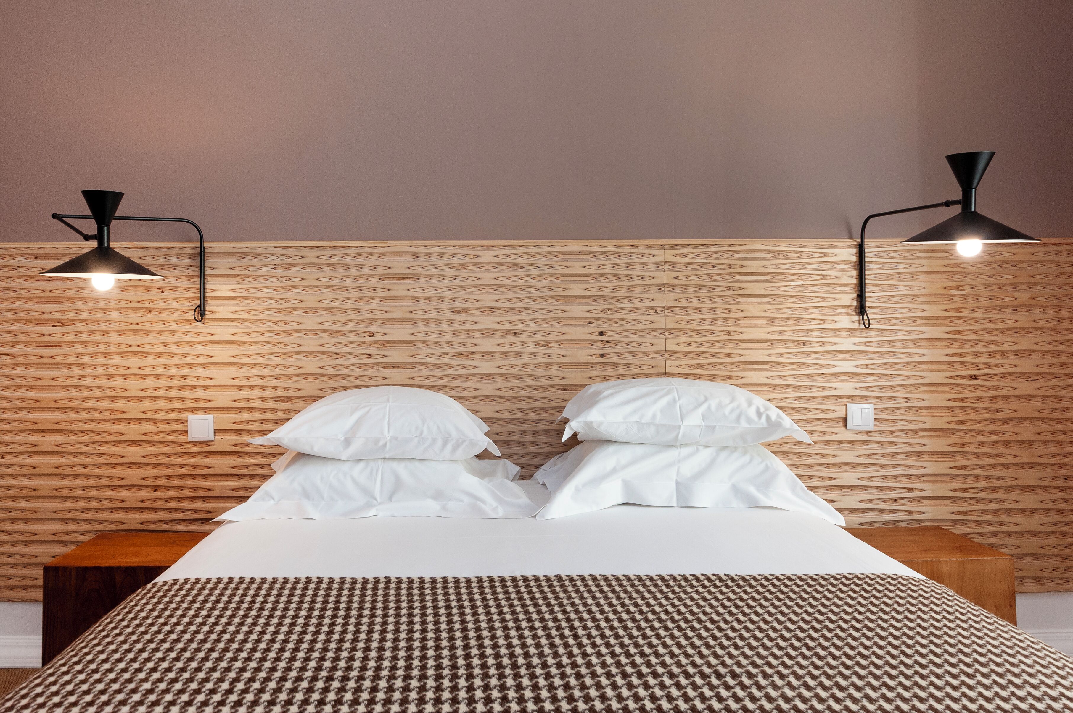 Ein ordentlich bezogenes Bett mit weißen Leinen und einer Bettdecke mit Muster, ein Holz-Kopfteil mit geometrischem Design und zwei schwarze, an der Wand befestigte Lampen auf jeder Seite.