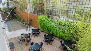 Un patio exterior con suelo de madera, mesas y sillas negras, y un jardín vertical cubierto de verde.