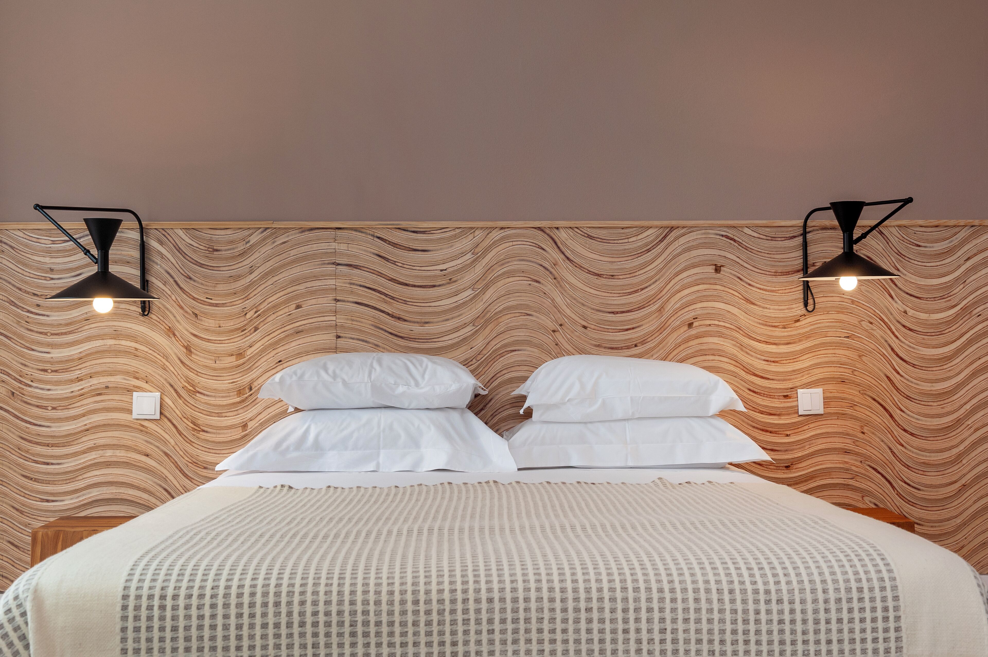 A bed with white linens, two black wall-mounted lights, and a wooden wavy-patterned headboard.