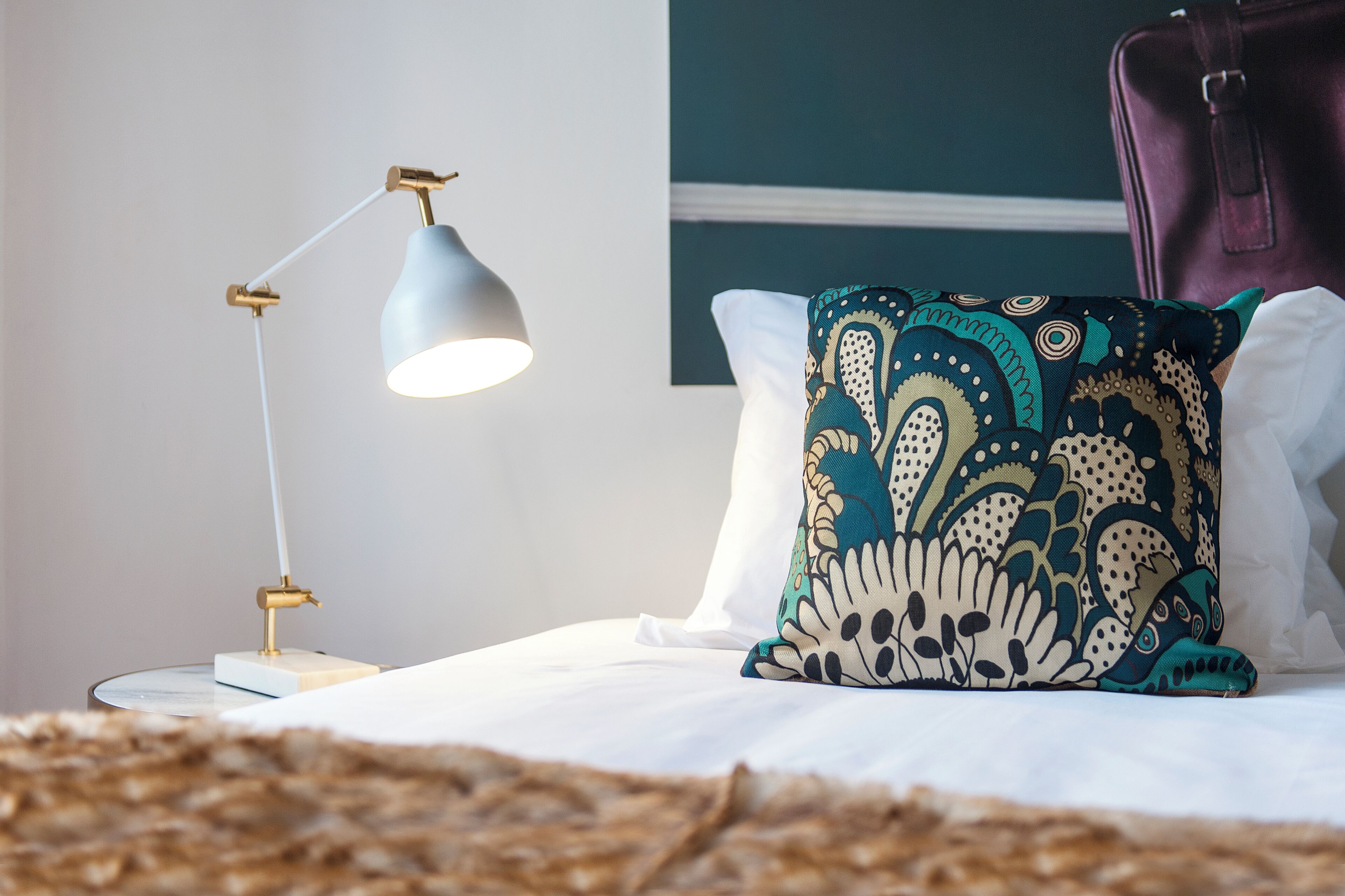 En seng med hvide sengetæpper, en pude med et mønster og en skrivebordslampe med hvid skærm.