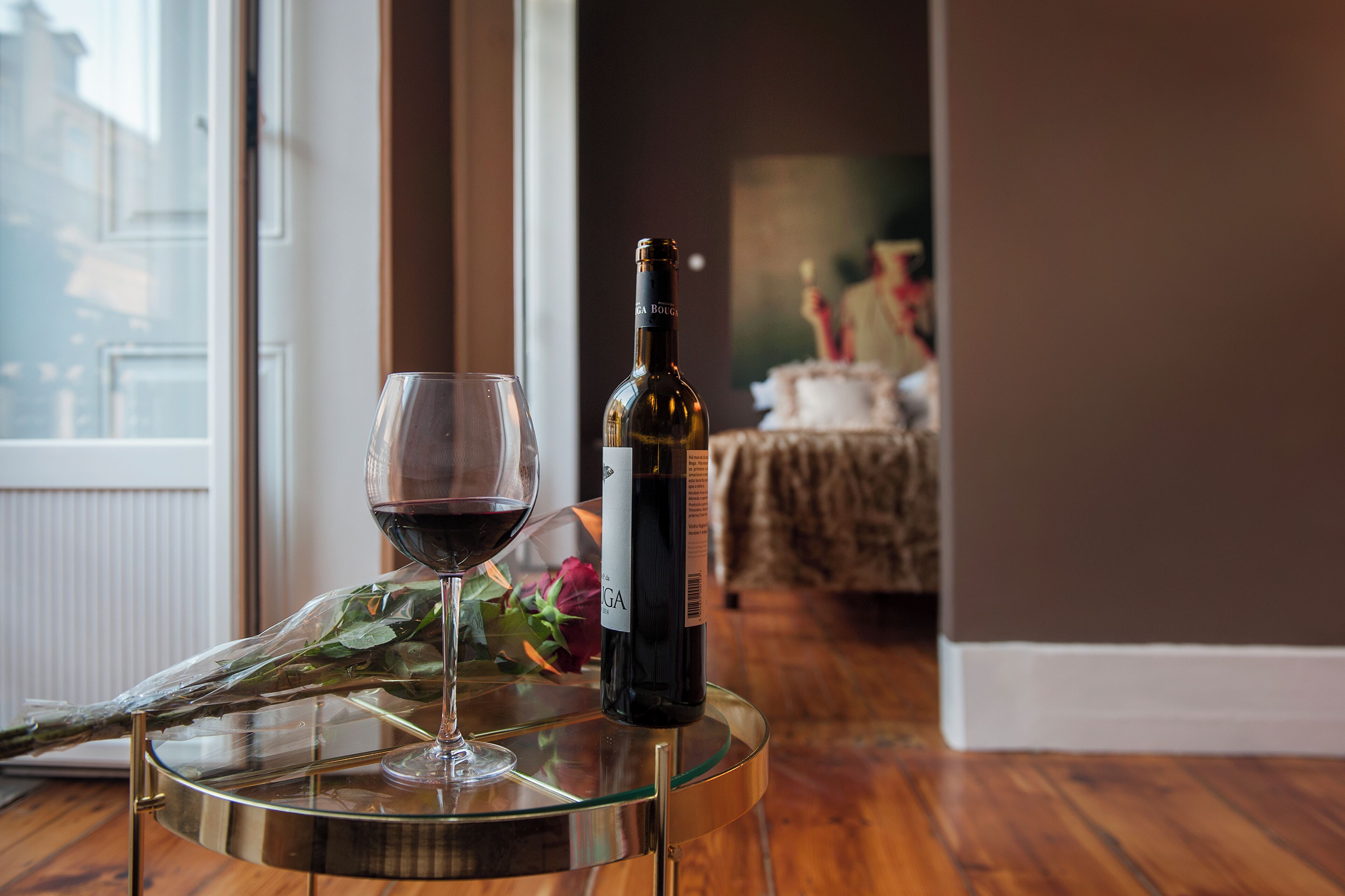 A glass of red wine and a bottle on a glass table with a view of a room in the background.