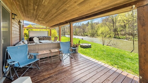 En overdækket terrasse med udendørs møbler, grill og udsigt til en flod og skov.