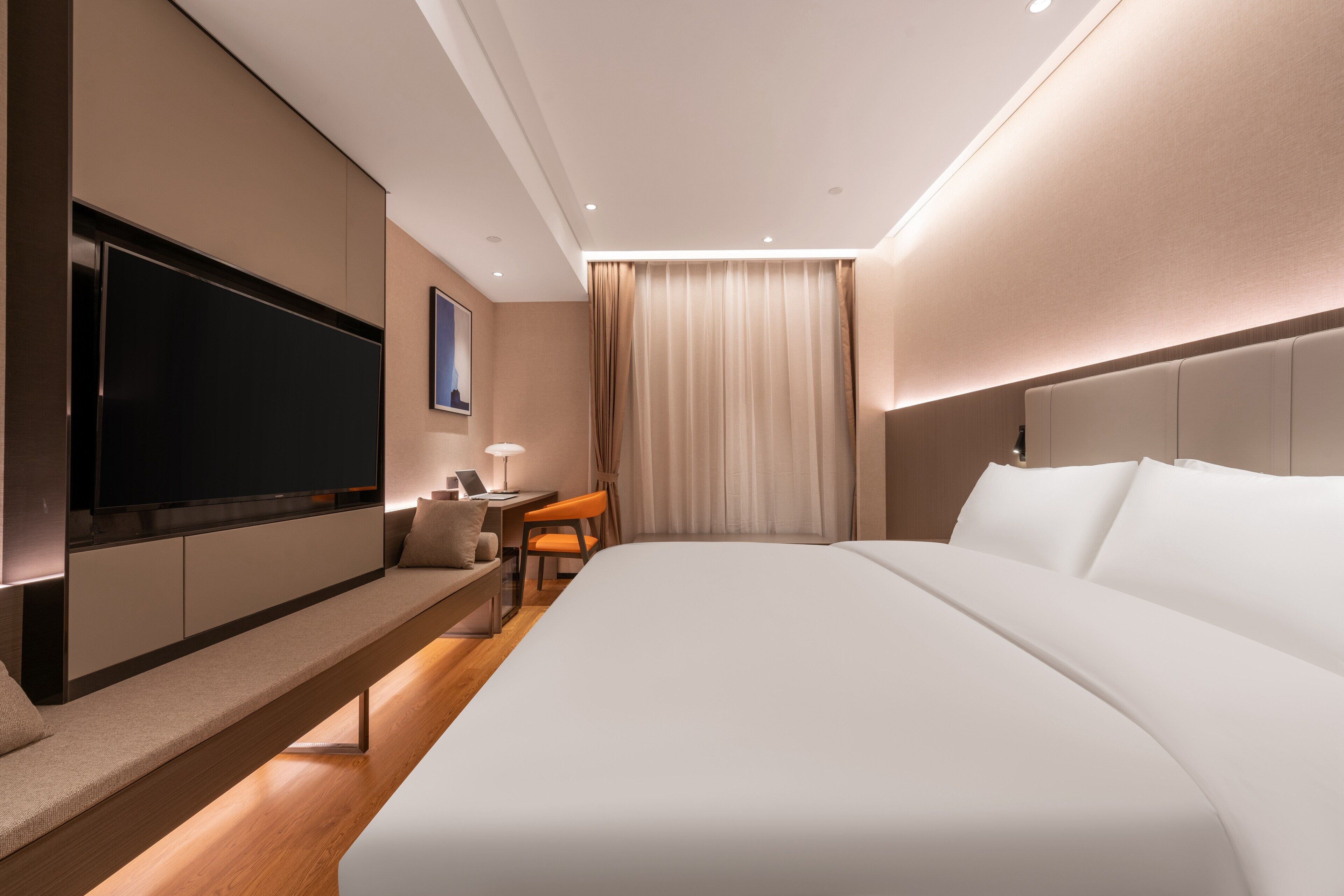 A modern hotel room with a large bed, a flat-screen TV mounted on the wall, a desk with a chair, and a window with curtains.