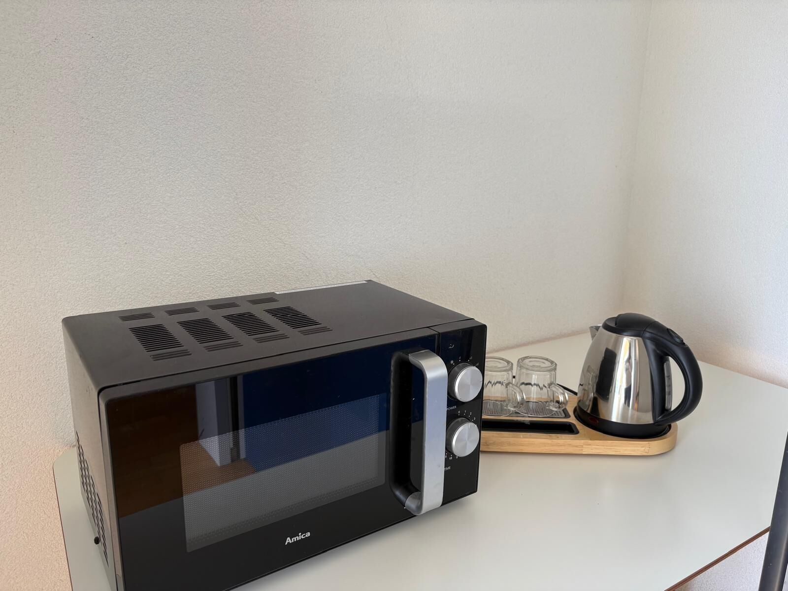 En svart mikrovågsugn från Amica med en digital display, en vattenkokare och ett fat med fyra glasmuggar, placerat på en vit bänkskiva.