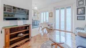 Un salon moderne doté d’un téléviseur, d’étagères en bois, d’un coin repas avec une table et des chaises, et de grandes fenêtres.