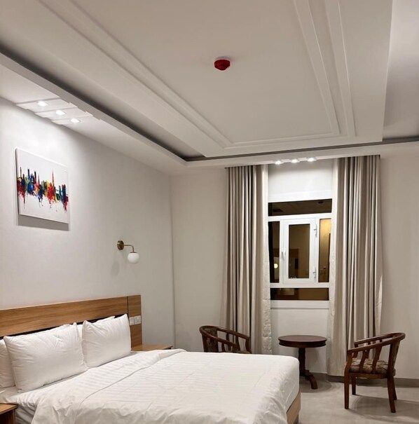 A modern hotel room with a large bed, two wooden chairs, a small table, and a painting on the wall. - 4 Guys Hotels (Muscat)