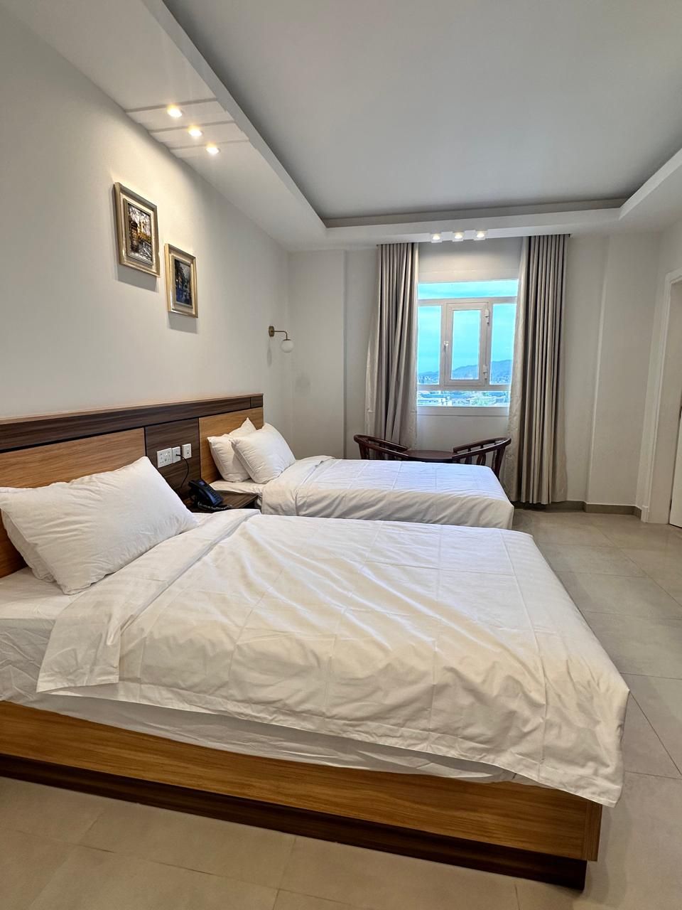 A hotel room with two beds, a wooden headboard, a nightstand, a chair, and a window with curtains.