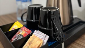 A coffee station with two black mugs, a stainless steel coffee pot, and various coffee packets. - 4 Guys Hotels (Muscat)