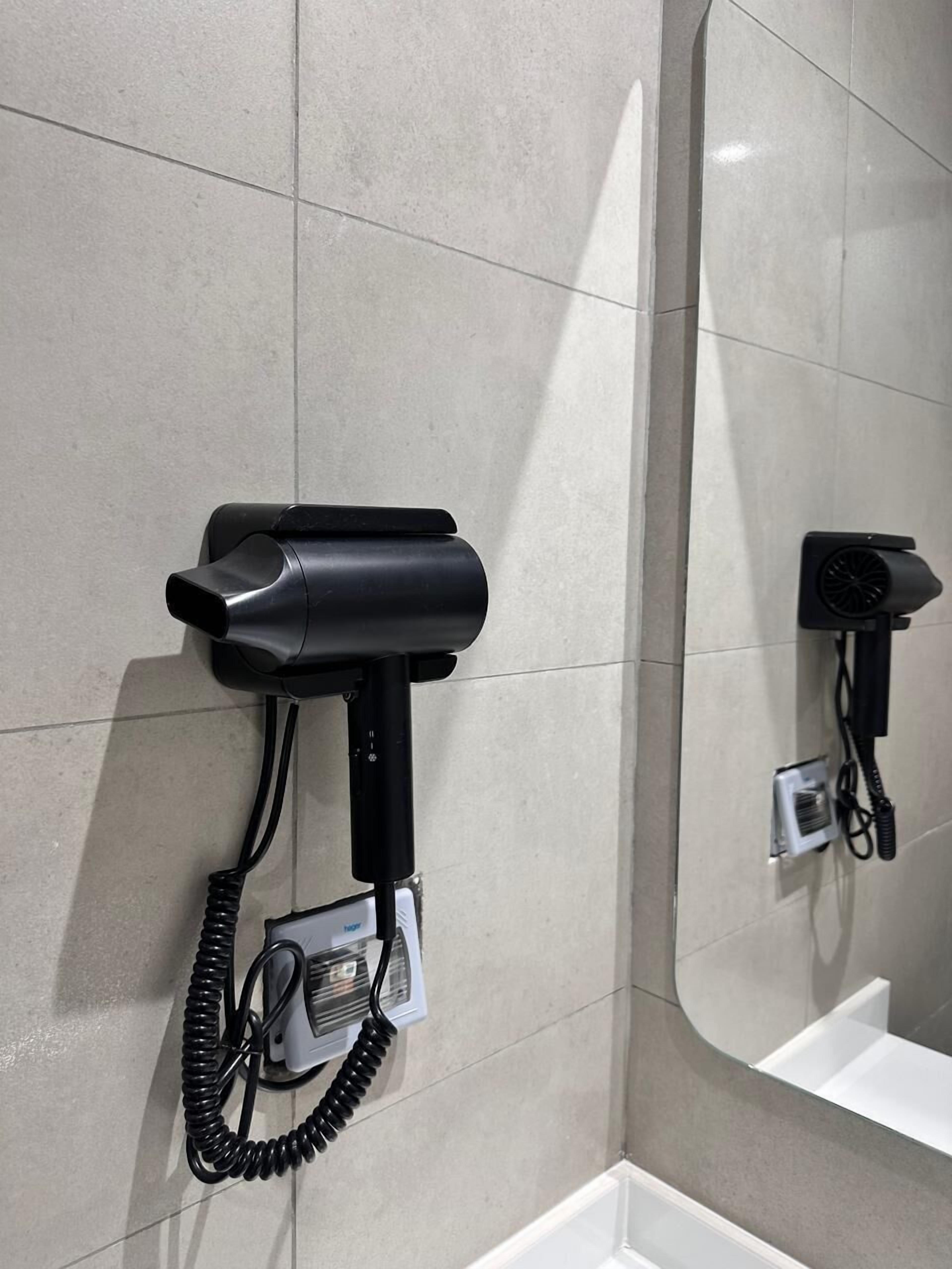 A bathroom with a hairdryer mounted on the wall, a small electrical outlet, and a mirror reflecting the hairdryer.