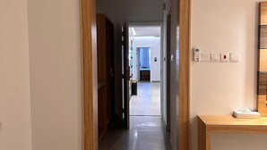 A hallway with a wooden door leading to a room with a bed and a blue curtain. There is a trash bin and a small table with a tissue box on the right. - 4 Guys Hotels (Muscat)
