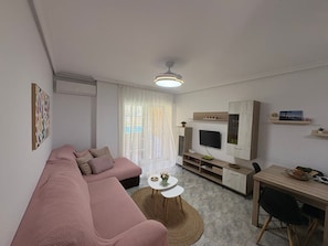 A modern living room with a pink sofa, a flat-screen TV, a dining area with a table and chairs, and a small rug.