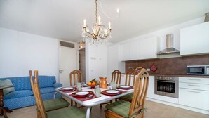 A dining area with a chandelier, wooden chairs with green cushions, a white table set for four, and a kitchen with white cabinets and a stainless steel oven. - GuestHost - Relax & Spacious Apartment X6 (Udine)