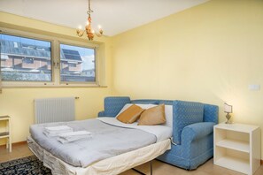 A bedroom with a bed, a blue sofa, a white nightstand, a chandelier, and a window with blinds. - GuestHost - Relaxing & Spacious Apartment for 6 (Udine)