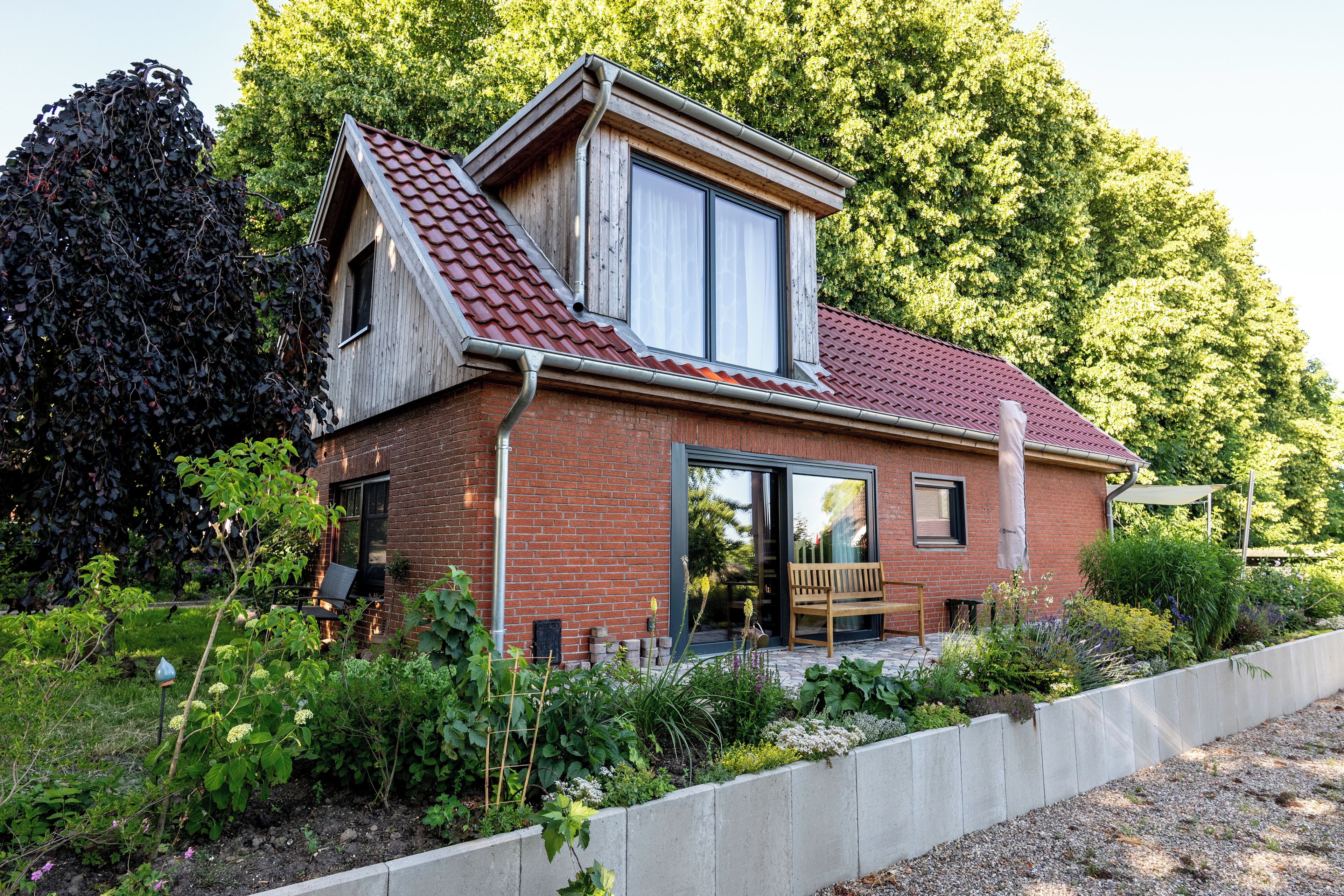 Une maison à deux étages, avec un toit recouvert de tuiles rouges, une grande fenêtre et un jardin où poussent différentes espèces de plantes.