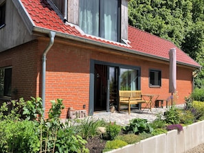 Une maison en briques avec un toit de tuiles rouges, un banc en bois et un jardin où poussent différentes espèces de plantes.