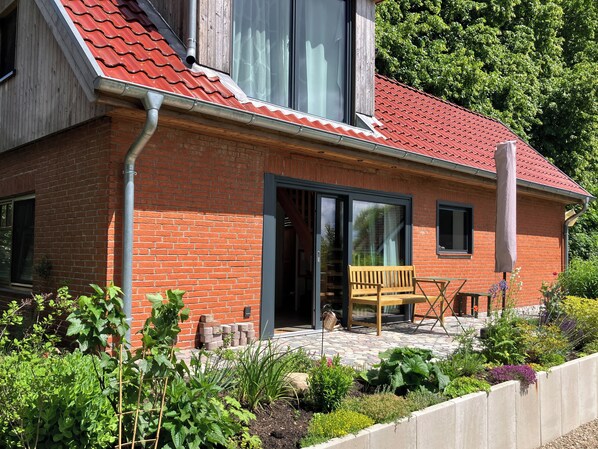 Une maison en briques avec un toit de tuiles rouges, un banc en bois et un jardin où poussent différentes espèces de plantes.