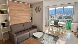 A modern living room with a sofa, a coffee table, and a dining area with chairs and a table. There is a large window with a view of a cityscape and mountains. - Newly renovated apartment 10 minutes from Collioure market. Ideal for families. (Collioure)