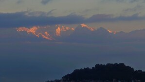 Ein Gebirge mit von Sonnenlicht erleuchteten Gipfeln, einem klaren Himmel und einer entfernten Hügelkette mit Gebäuden.