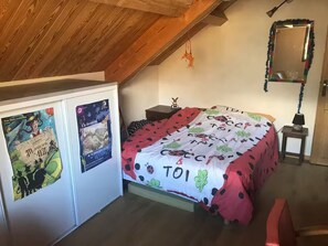 Ein Schlafzimmer mit schrägen Wänden, einem Bett mit Kissenüberwurf in Punkten und Fruchtmuster, einem kleinen Nachttisch mit Lampe sowie Postern an der Wand.