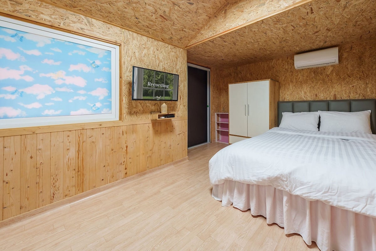 Quarto com painéis de madeira, uma cama, televisão e uma porta que dá para outro espaço.