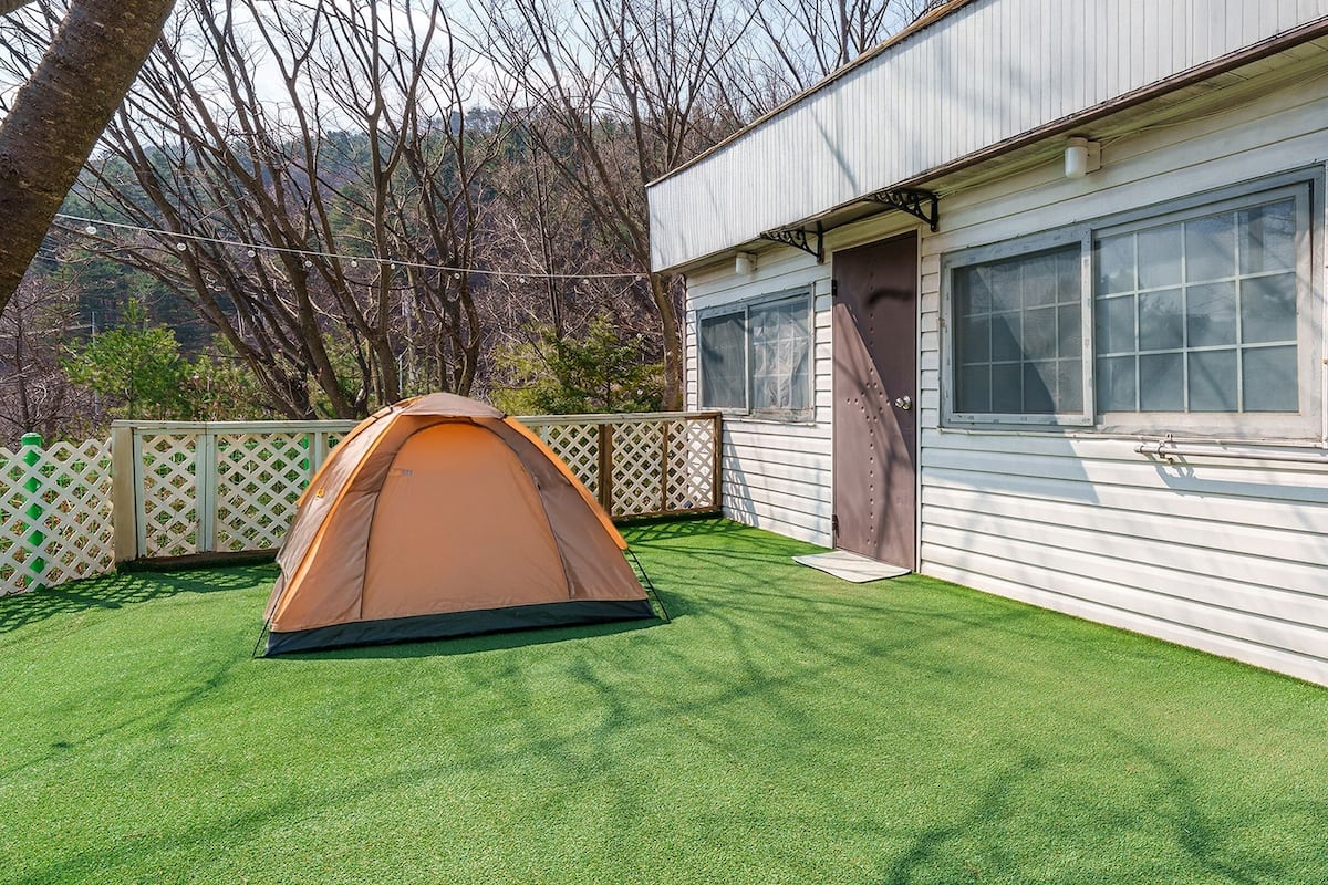 Tenda de campismo montada em relva artificial, em frente a uma casa móvel com janela e porta.