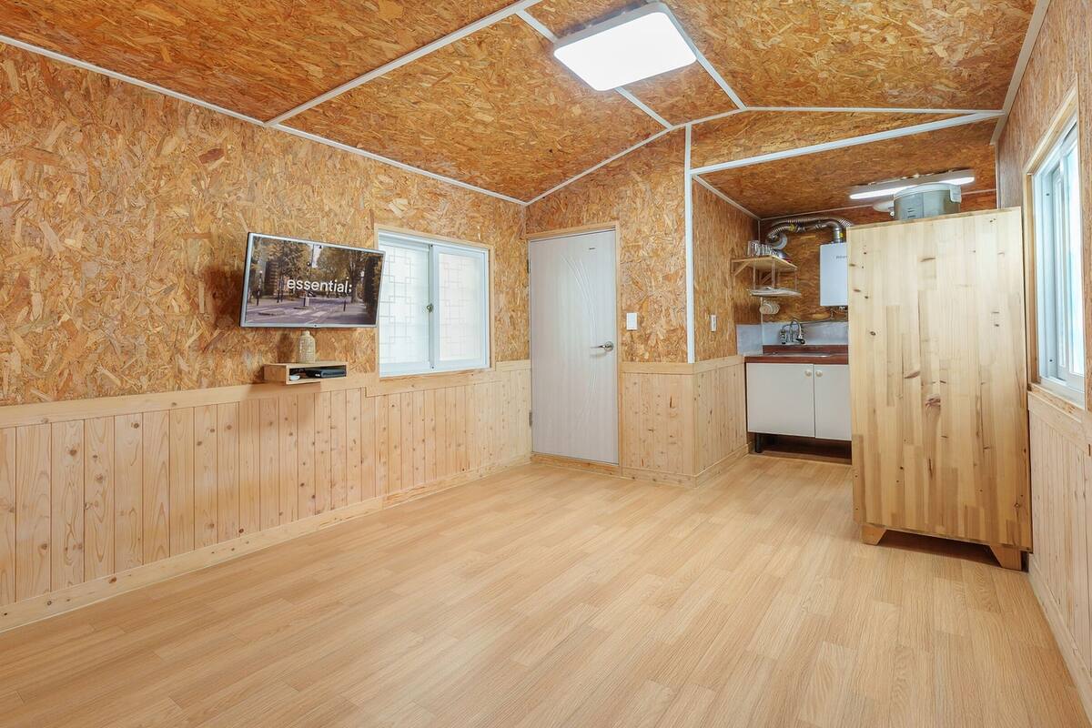 Quarto com paredes e chão de madeira, televisão montada na parede, uma janela e uma porta.