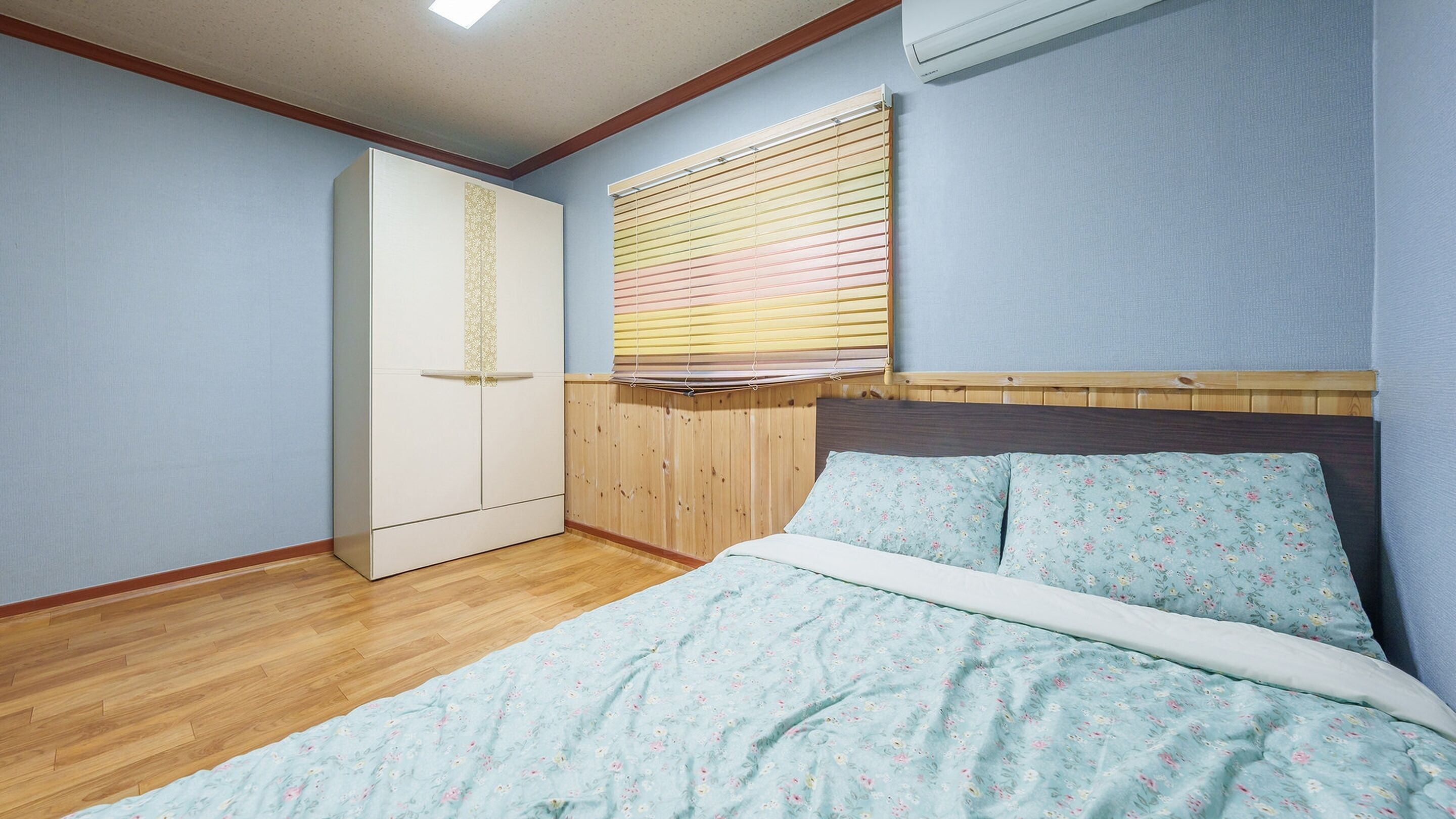 A bedroom with a wooden headboard, a bed with floral bedding, a wardrobe, and a window with blinds.