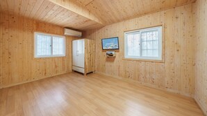 Una habitación con paredes y suelos de madera, un armario blanco, un televisor de pantalla plana montado en la pared y dos ventanas con persianas de diseño bambú.