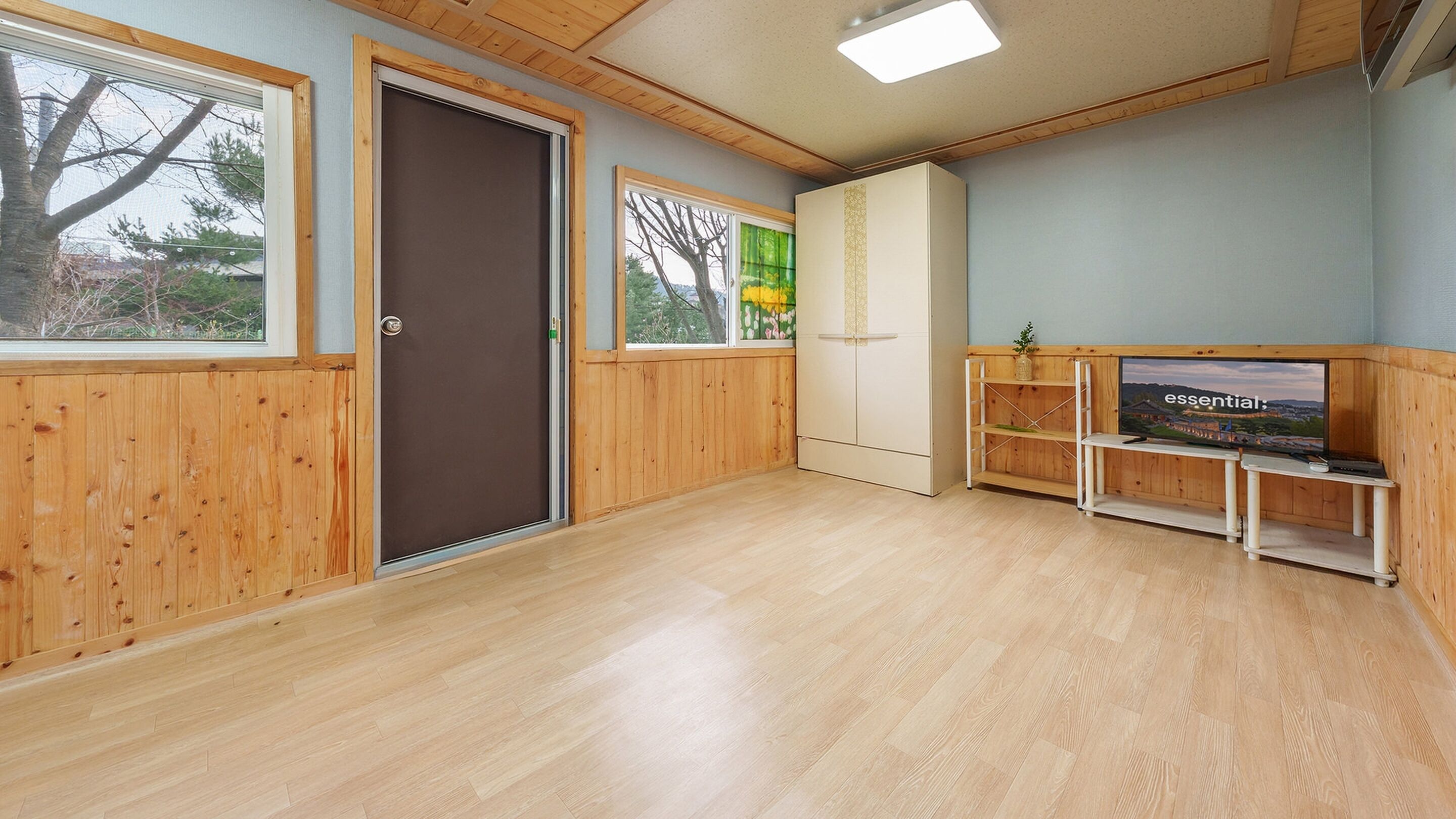 Ein Zimmer mit Holzwänden, einer Tür, einem Fenster mit Blick, einem Kleiderschrank und einem Fernsehschrank mit Fernseher.