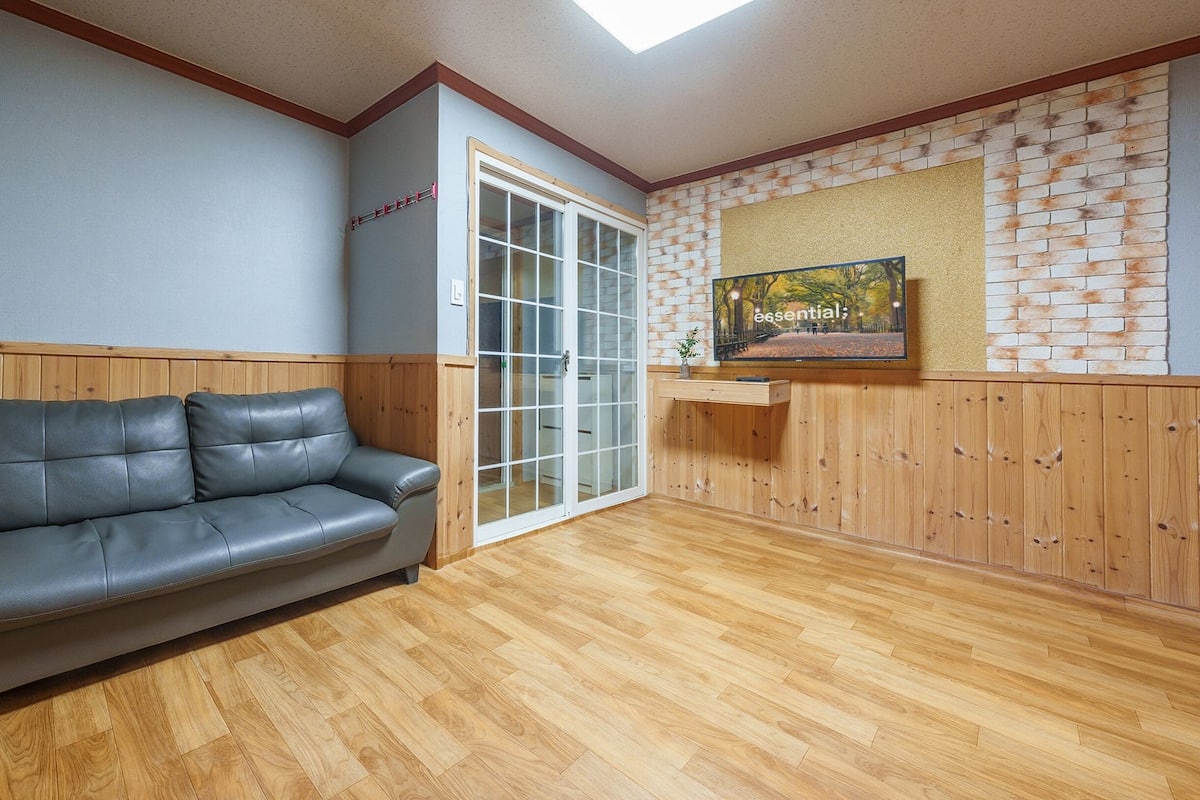 Quarto com piso de madeira, sofá de couro cinza, porta de vidro deslizante e televisão na parede exibindo 'essencial'.