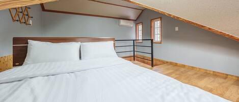 Un dormitorio con cabecera de madera, cama con ropa de cama blanca, suelo de madera y techo inclinado con vigas a la vista.
