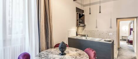 Habitación de hotel moderna con una mesa redonda de mármol, sillas moradas, un pequeño objeto decorativo, una zona de cocina y vistas a la habitación contigua.