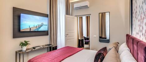 A modern hotel room with a large bed, a TV mounted on the wall, a desk with a plant, and a view of the sea through the window.