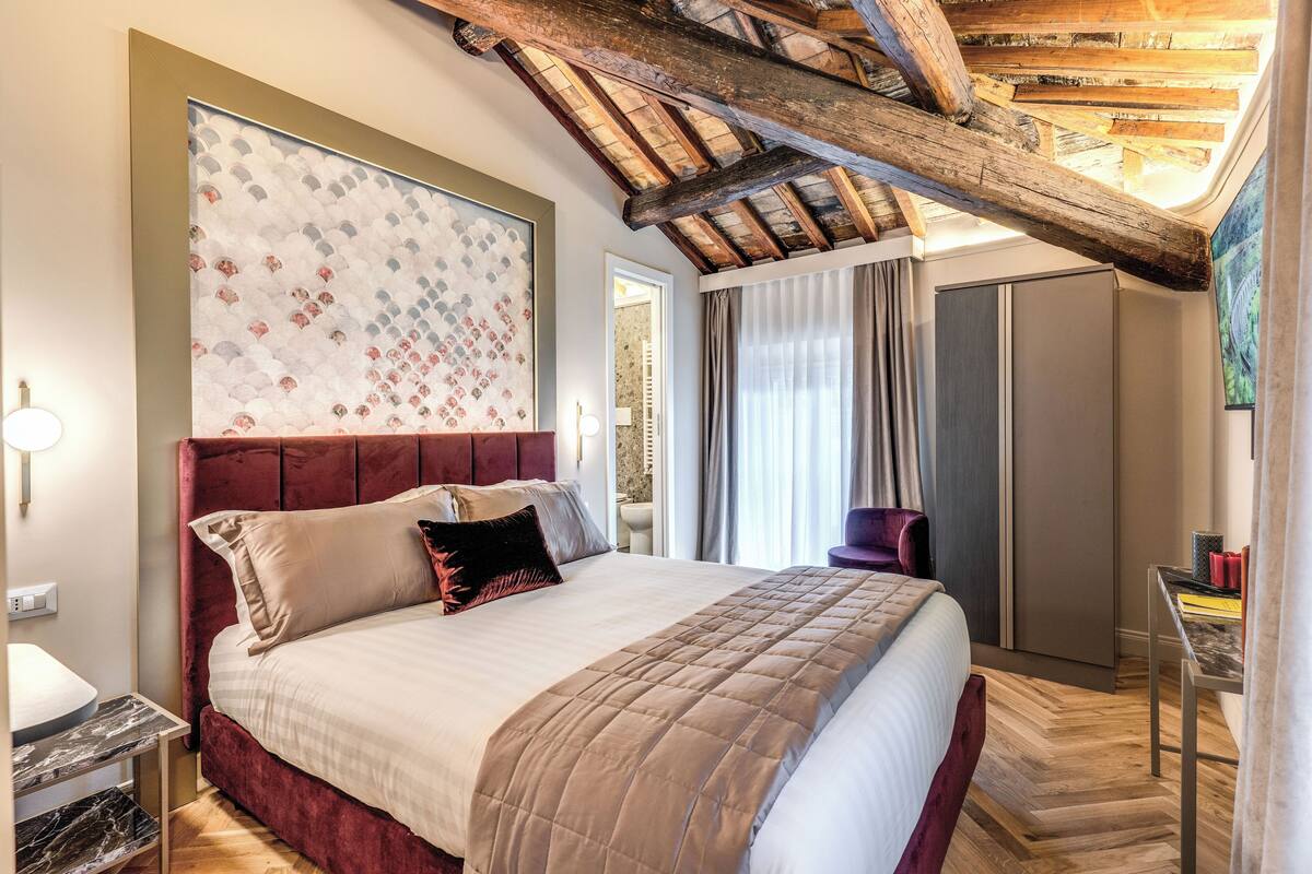 A spacious bedroom with a large bed, wooden ceiling beams, a modern bathroom, and a view of the outdoors.