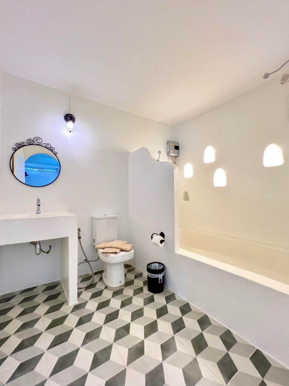 Un baño con lavamanos, inodoro y bañera blancos. Hay un espejo con un marco decorativo, una lámpara de pared y un cubo de basura.