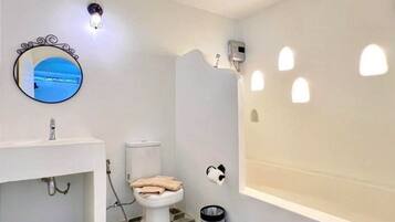 Un baño con lavamanos, inodoro y bañera blancos. Hay un espejo con un marco decorativo, una lámpara de pared y un cubo de basura.