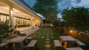 An outdoor patio area with stone tables and chairs, a checkered lawn, and a building with large windows and a covered seating area. - Villa La Flora Kaeng Krachan (Kaeng Krachan)