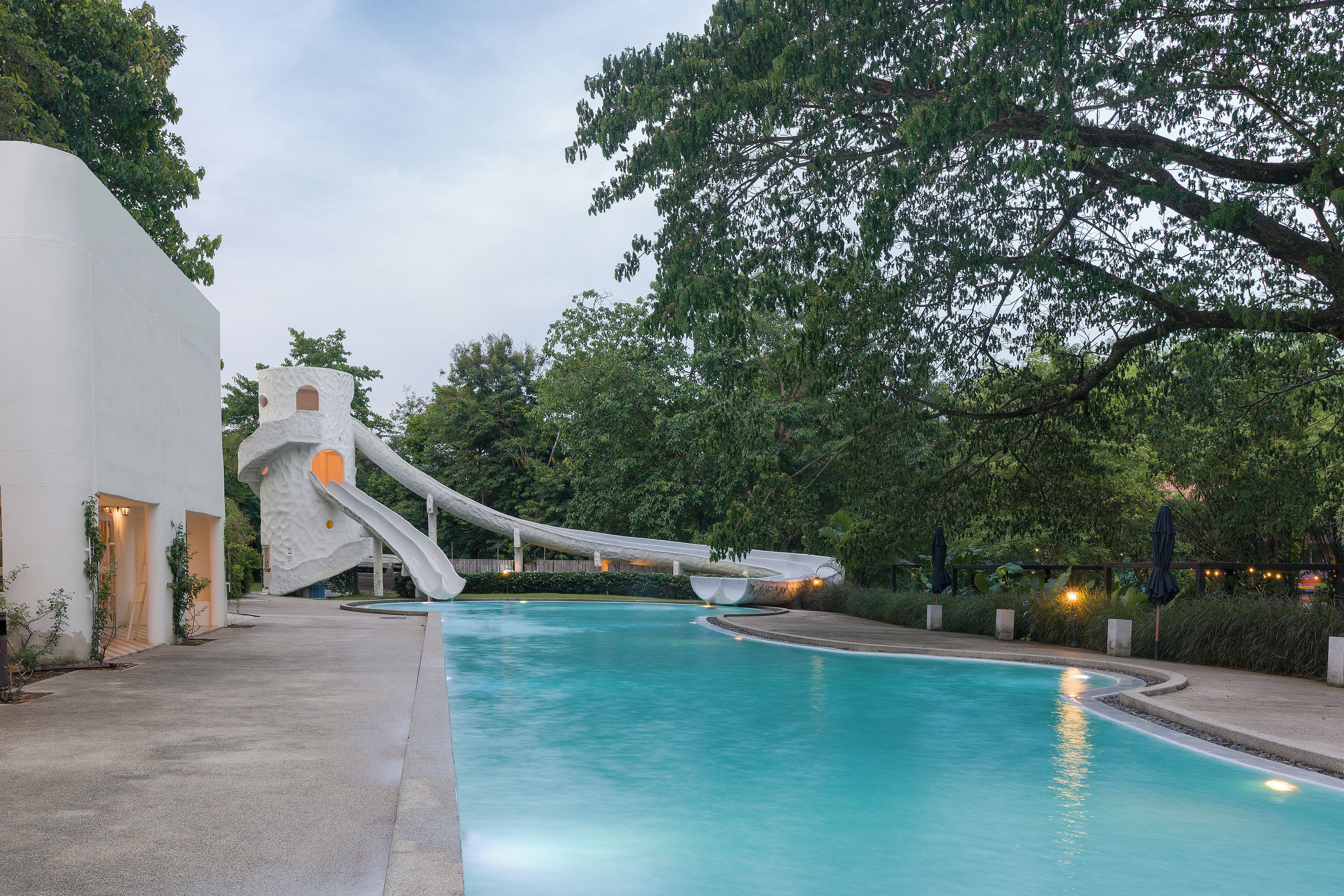 A swimming pool with a water slide, surrounded by trees and a white building with a unique architectural design.