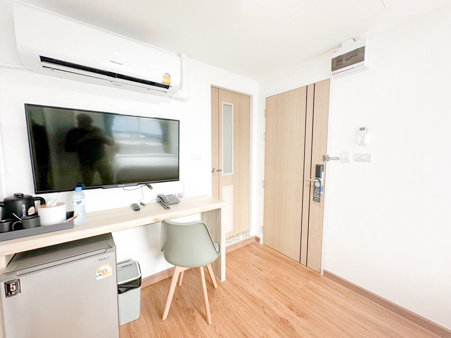 A compact hotel room with a kitchenette, a flat-screen TV, a small refrigerator, a desk with a chair, and a wooden wardrobe.