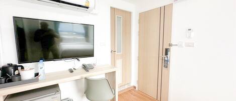 A compact hotel room with a kitchenette, a flat-screen TV, a small refrigerator, a desk with a chair, and a wooden wardrobe.