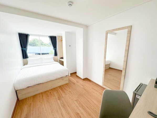 A hotel room with a bed, a desk, a chair, a mirror, and a window with curtains. - H-Banaat Hotel (Bangkok)