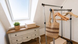 Ein Zimmer mit Fenster, einem Holzschrank, einer HäkelTasche auf einem Metallgestell und einer kleinen Pflanze.