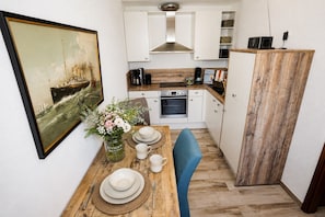 Cozinha compacta com armários brancos, uma mesa de jantar de madeira com pratos e xícaras, um vaso de flores e um quadro na parede.